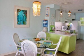 Un espace repas avec une table en verre, des chaises blanches et un îlot de cuisine vert équipé d’appareils électroménagers.