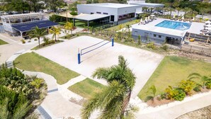 Een sportveld met een volleybalnet, omringd door een aangelegde tuin met palmbomen en een zwembad.