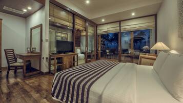 Quarto de hotel com uma cama grande, uma mesa de trabalho com televisão, uma cadeira e vista para palmeiras pela janela.