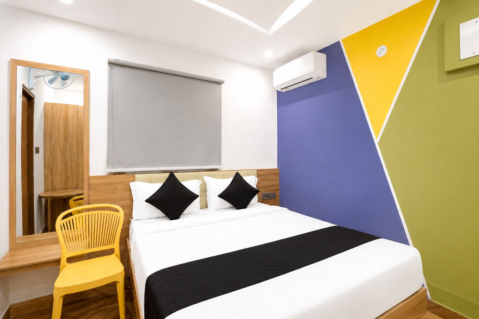 A hotel room with a bed, a desk with a chair, an air conditioning unit, and a mirror.