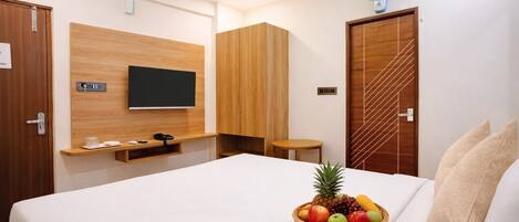 A modern hotel room with a large bed, a TV mounted on the wall, a wooden wardrobe, a small table, and a fruit basket on the bed.