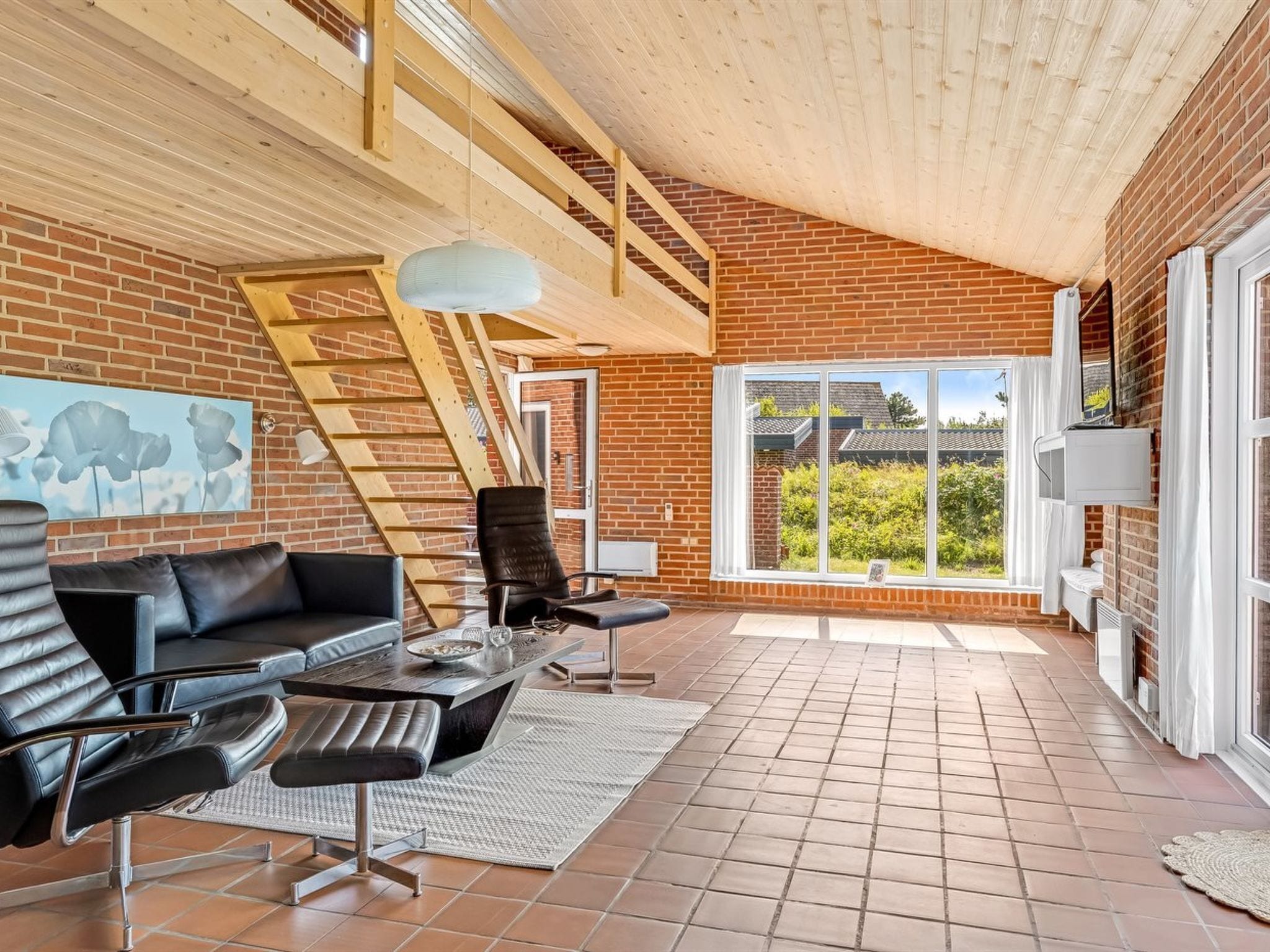 A spacious living area with a brick wall, a wooden ceiling, a black leather sofa, a coffee table, and a large window offering a view of greenery.