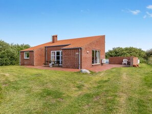 Una casa de ladrillo de un piso con techo de tejas, una chimenea visible y una estructura pequeña adosada con una mesa y sillas.