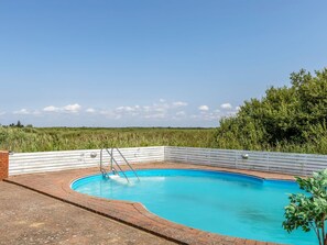 Una piscina con cubierta de madera y escalera metálica, rodeada de follaje y un cielo despejado.