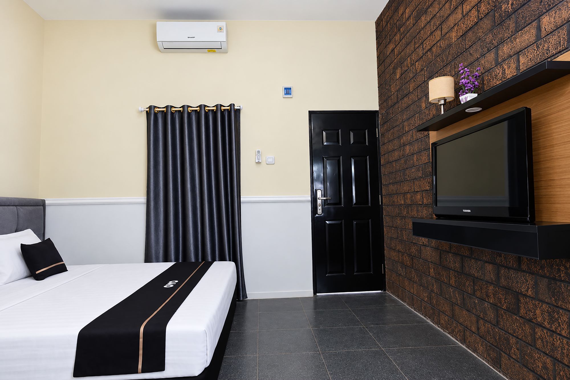 A hotel room with a bed, a black door, a mounted TV, and a wall-mounted air conditioner.