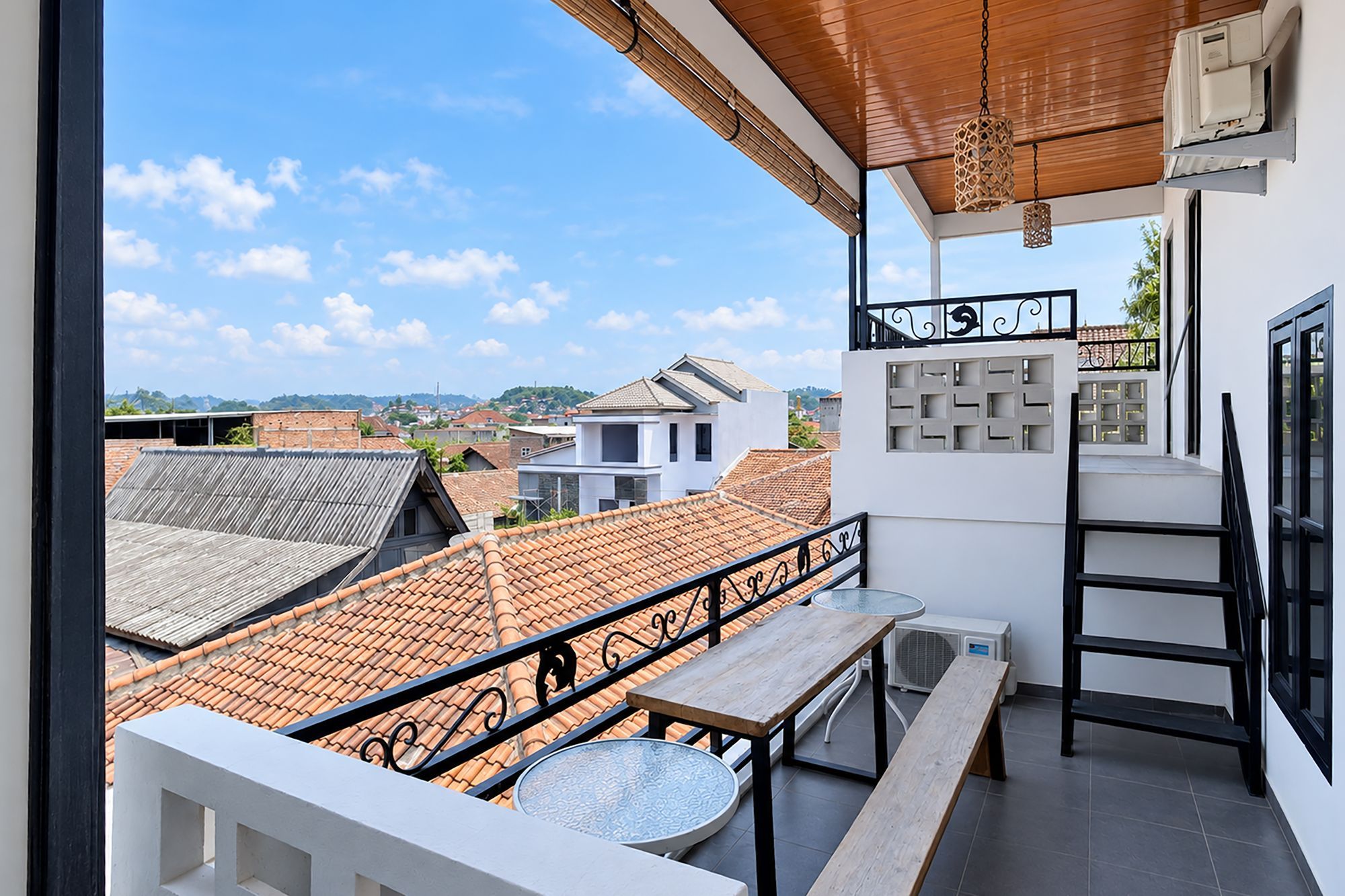 A balcony with a view of the cityscape, a wooden table, a metal railing, and a white wall with a decorative panel.