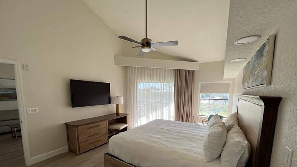 Ein Schlafzimmer mit einem Bett, einem Flachbildfernseher, einem Deckenventilator und einem Vorhangfenster.
