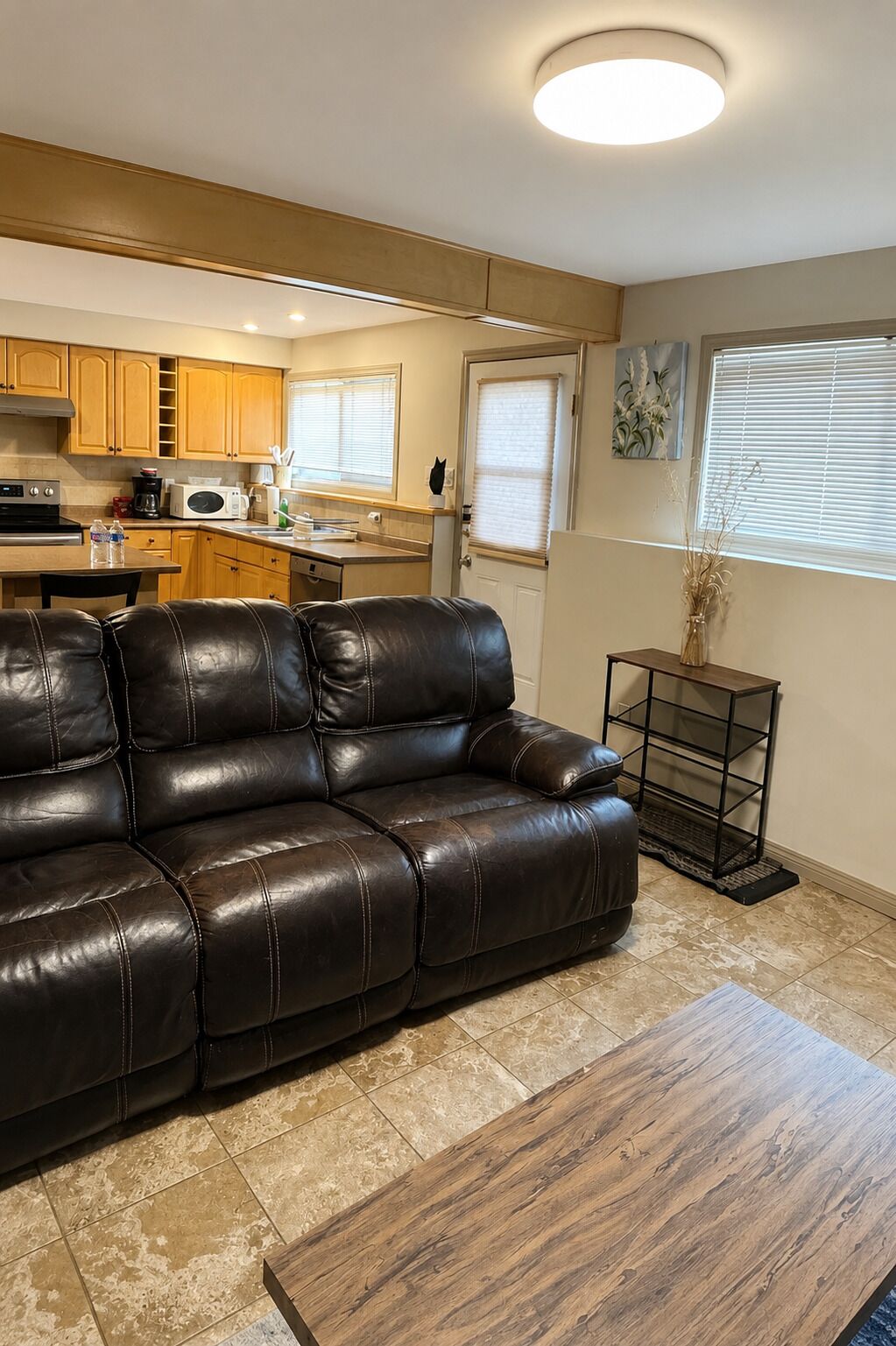 A living room with a leather sofa, a wooden coffee table, a kitchen area with wooden cabinets, a microwave, and a sink.