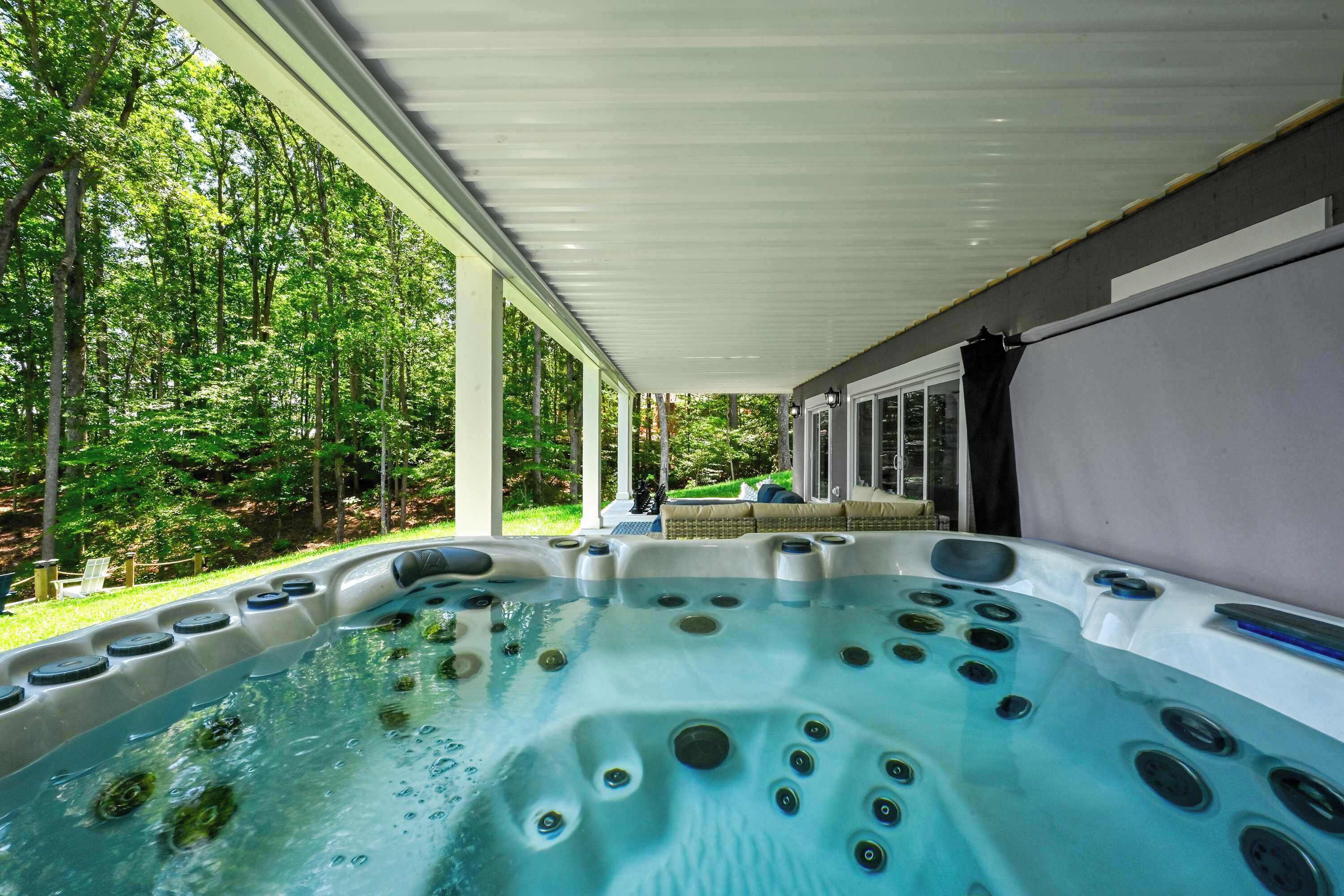 Un jacuzzi con múltiples chorros, rodeado por un porche cubierto con vista a un bosque verde exuberante.