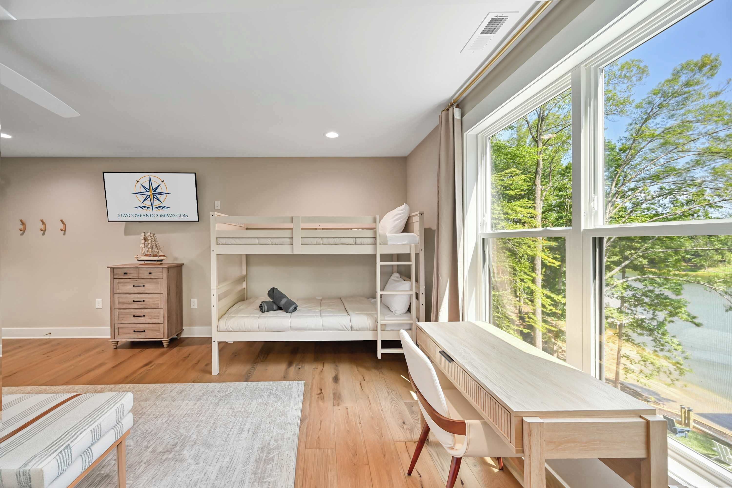 A bedroom with a bunk bed, a wooden desk, a dresser, and a large window offering a view of trees.