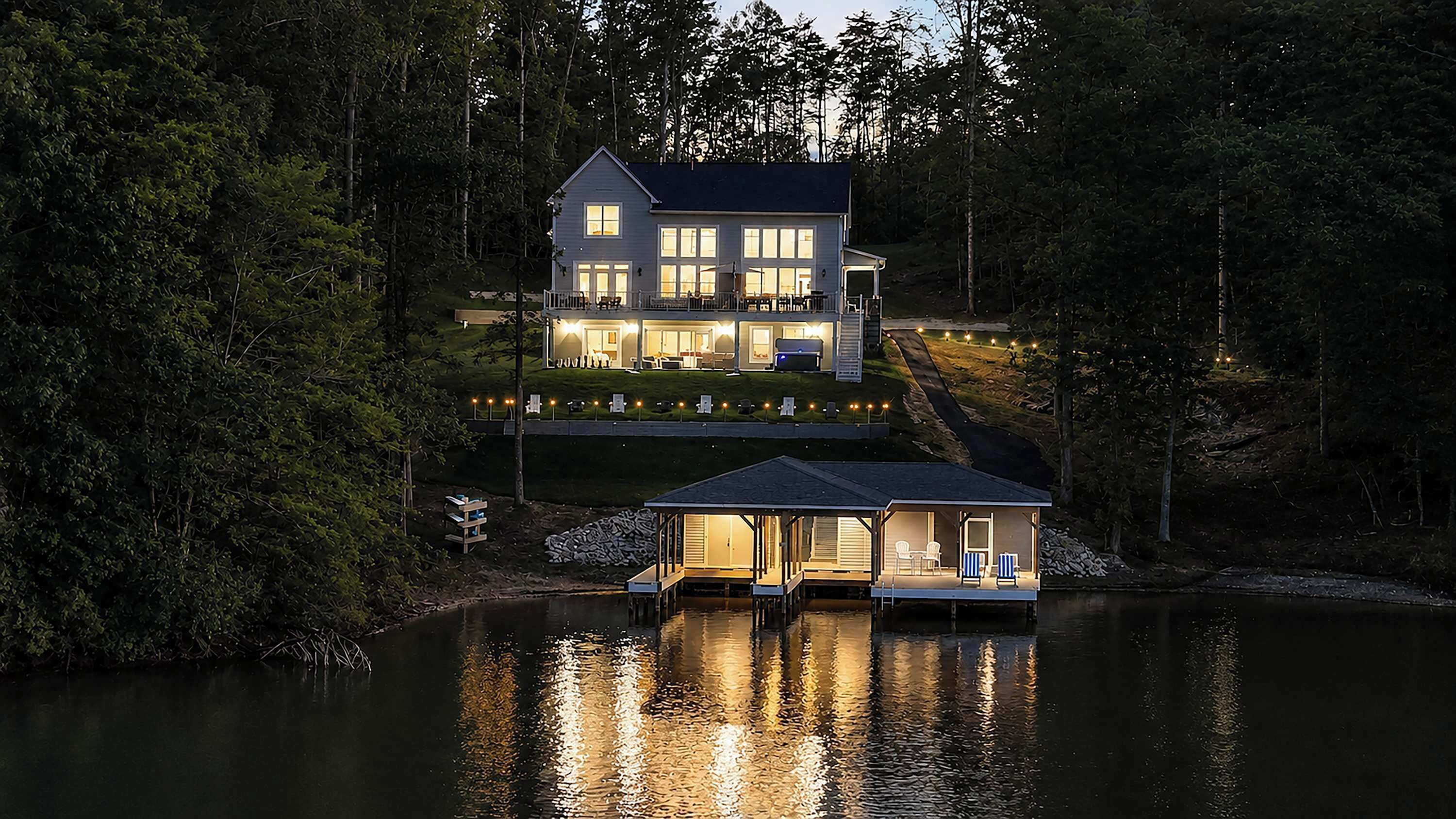 A two-story house with a dock extending into a lake, surrounded by dense forest.