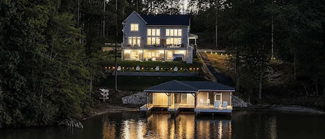 A two-story house with a dock extending into a lake, surrounded by dense forest.