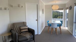 Ein modernes Wohnzimmer mit einer Couch, einem kleinen Tisch, einem Essbereich mit Stühlen und einem großen Fenster mit Blick auf Grünflächen.