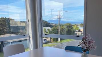 A room with a white table and a vase of flowers, large windows with a view of a residential area and a body of water.