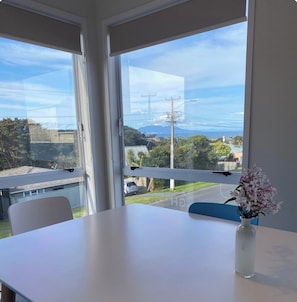 Ein Zimmer mit weißem Tisch und Blumen in einer Vase, große Fenster mit Blick auf einen Wohnbereich und ein Gewässer.
