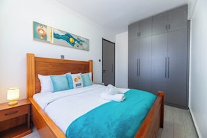 A neatly made bed with a wooden headboard, a nightstand with a lamp, and a grey wardrobe with two doors. - Cozy 2 BR with Heater,Gym,Pool,ample parking on 3floors at Lavington,Nairobi (Nairobi)