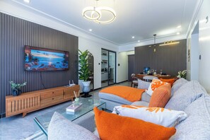 Ein modernes Wohnzimmer mit Flachbildfernseher, einer Couch mit orangefarbenen Kissen, einem Glastisch und einer Holzkonsole.