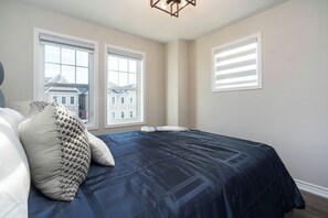 A bedroom with a bed, two windows, and a view of houses outside.