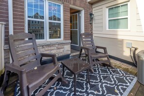 A patio area with two wooden chairs and a small table, a patterned rug, and a brick wall with windows.