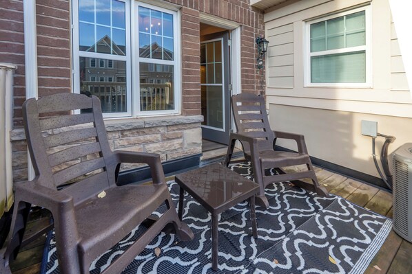 A patio area with two wooden chairs and a small table, a patterned rug, and a brick wall with windows.