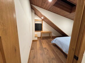 Ein kleiner Raum mit schrägem Dach, einer Holzbänke, einem Bett und einem Fenster.