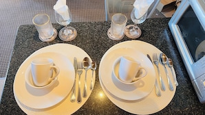 Une table à manger est dressée avec deux assiettes, des tasses, des couverts et des serviettes, ainsi qu’un verre d’eau et un verre de vin posés sur des sous-verres.
