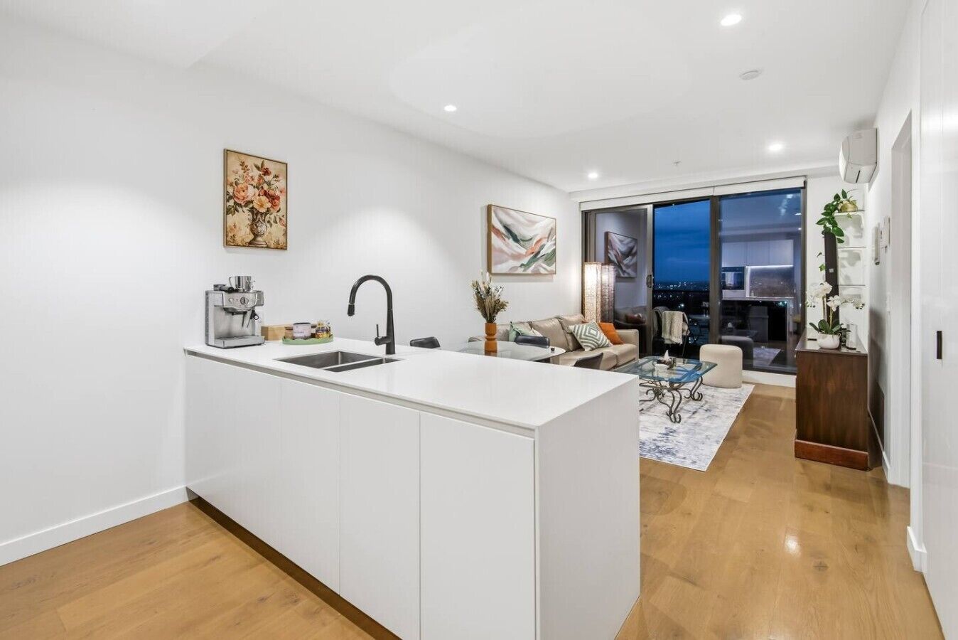 Een moderne keuken met een centraal eiland, een eethoek met een glazen tafel, en een woonkamer met een bank en een groot raam.