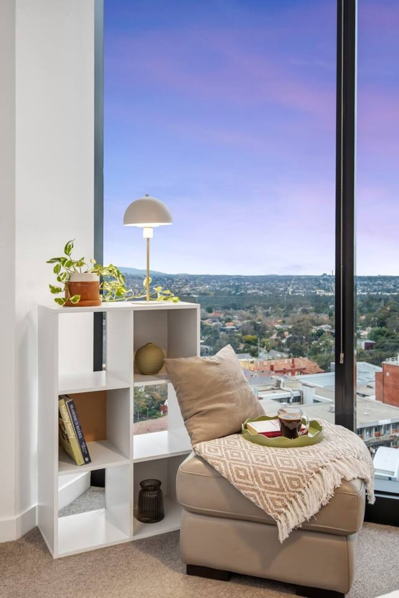 A modern living room with a large window, a white bookshelf, a beige sofa, and a table with a lamp.