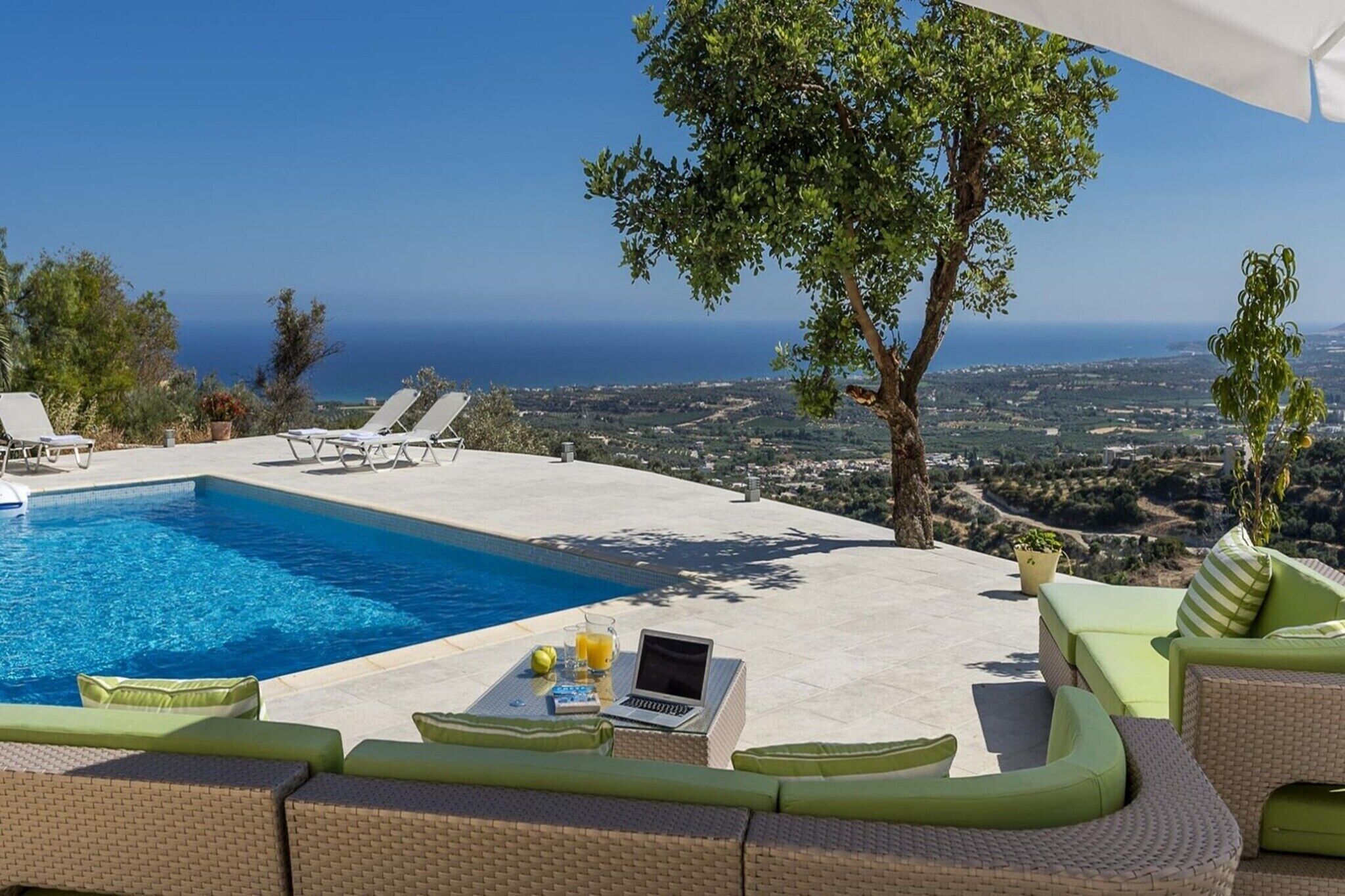 A poolside area with a view of the sea, lounge chairs, and a table with a laptop and drinks.