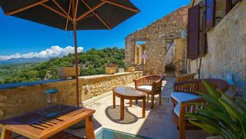 A terrace with wooden furniture, a table, and an umbrella, overlooking a mountainous landscape.