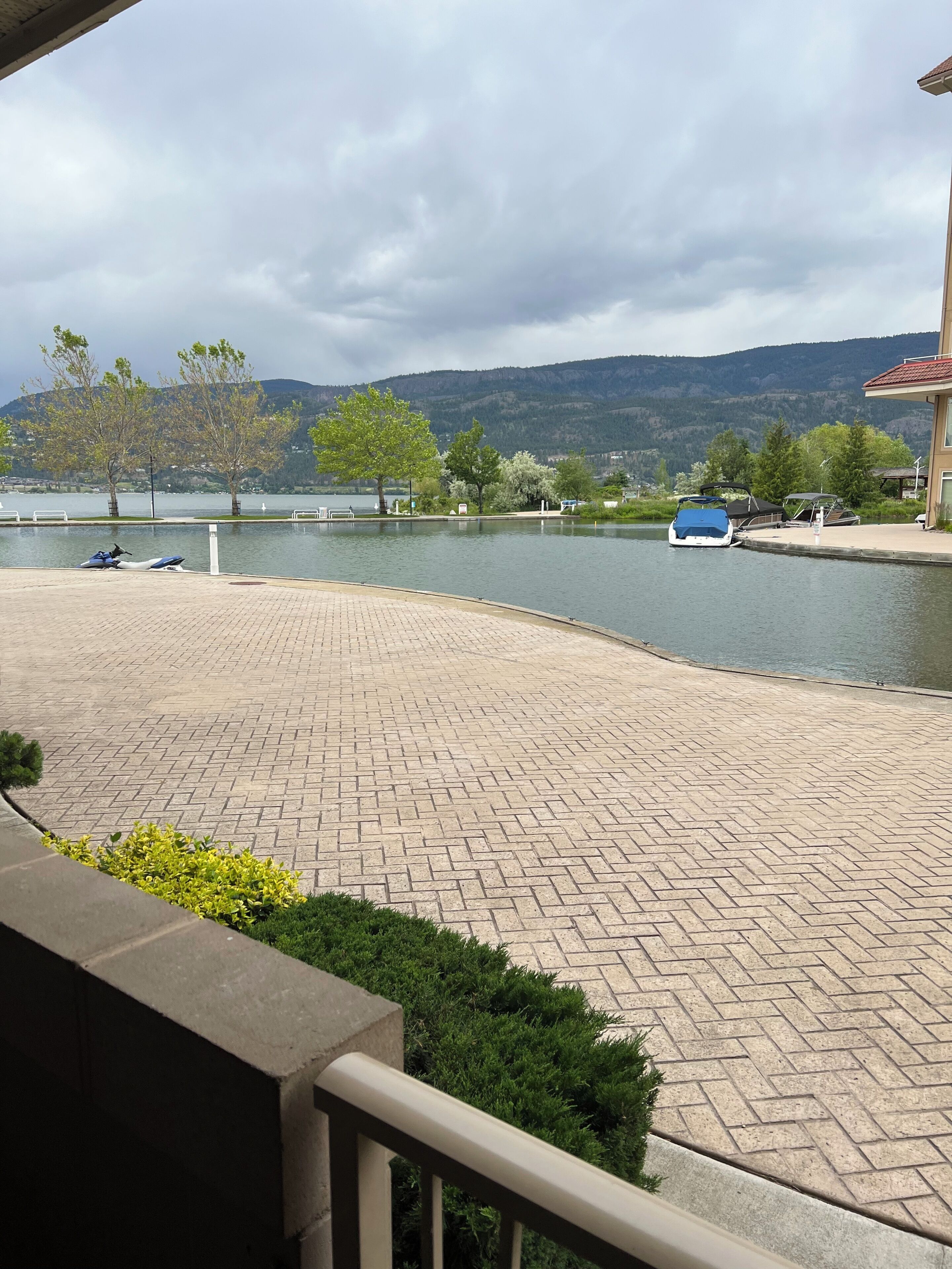 A paved waterfront area with boats docked, trees, and a view of hills in the background.