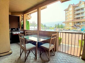 An outdoor patio with a glass table and chairs, a barbecue grill, and a view of a body of water and buildings. - Sunset Waterfront Resort, Executive Townhouse 113. Luxury Lakeside, 3 Bed/2 Bath (Kelowna)