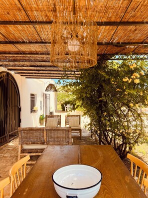 Ein Essbereich im Freien mit einem Holztisch und Stühlen, einer hängenden Leuchte und Blick auf einen Garten mit Bäumen und einem Gebäude im Hintergrund.