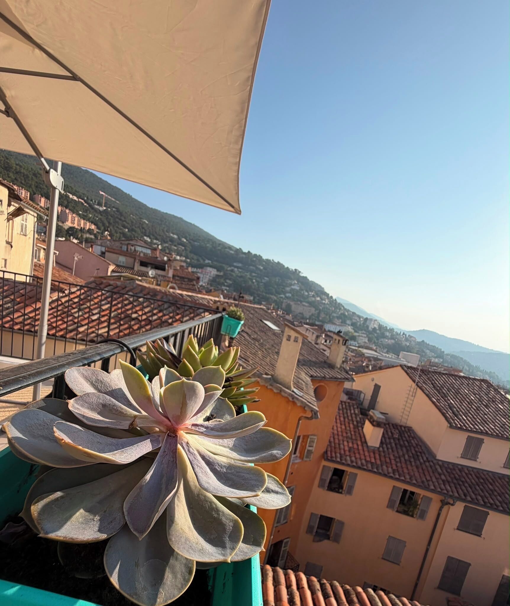 Un balcon donnant sur une ville située sur une colline, une grande plante et un auvent de couleur beige.
