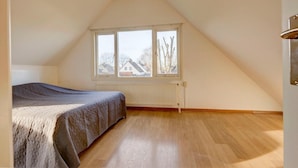 A bedroom with a sloped ceiling, a bed, a radiator, and a window with a view of houses. - Holiday Home in South Holland (Noordwijkerhout)