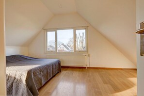 A bedroom with a sloped ceiling, a bed, a radiator, and a window with a view of houses. - Holiday Home in South Holland (Noordwijkerhout)