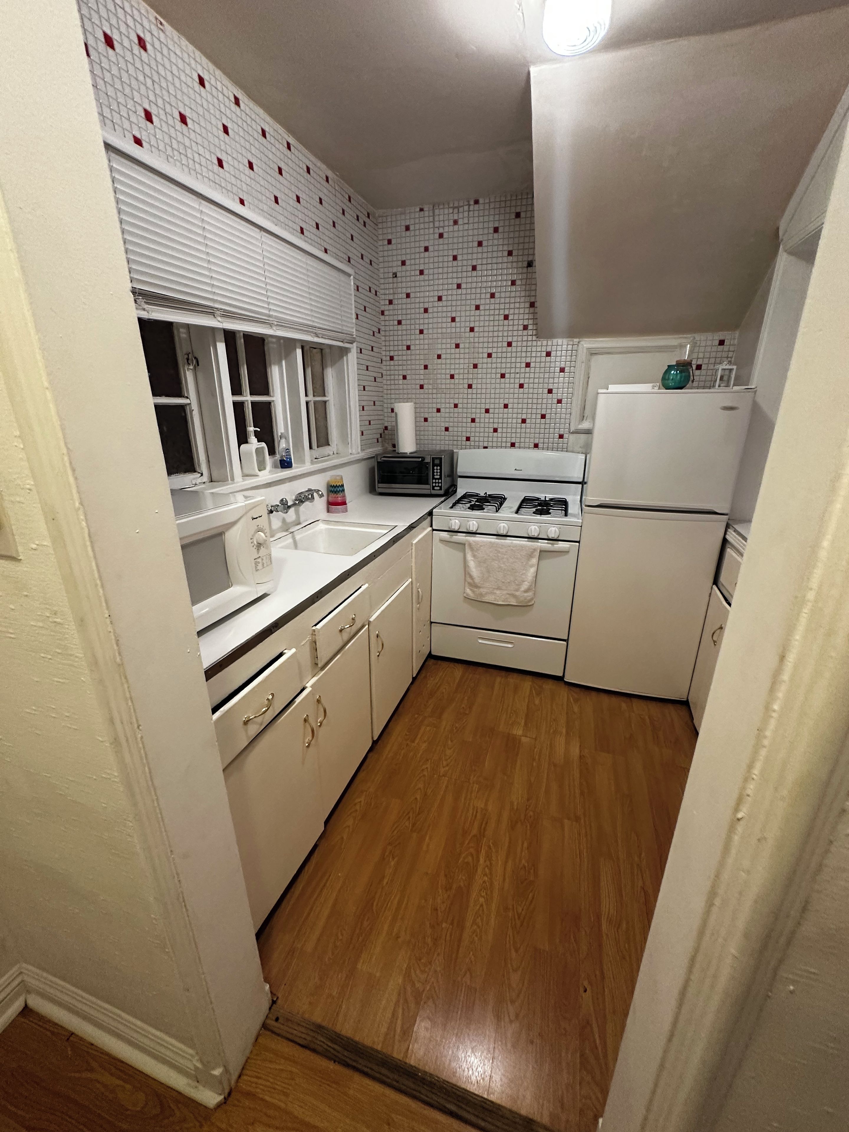 A compact kitchen with a gas stove, a built-in oven, a refrigerator, and a microwave. There is a window with blinds and a sink with a faucet.