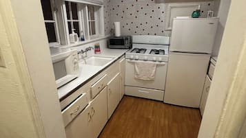 A compact kitchen with a gas stove, a built-in oven, a refrigerator, and a microwave. There is a window with blinds and a sink with a faucet.