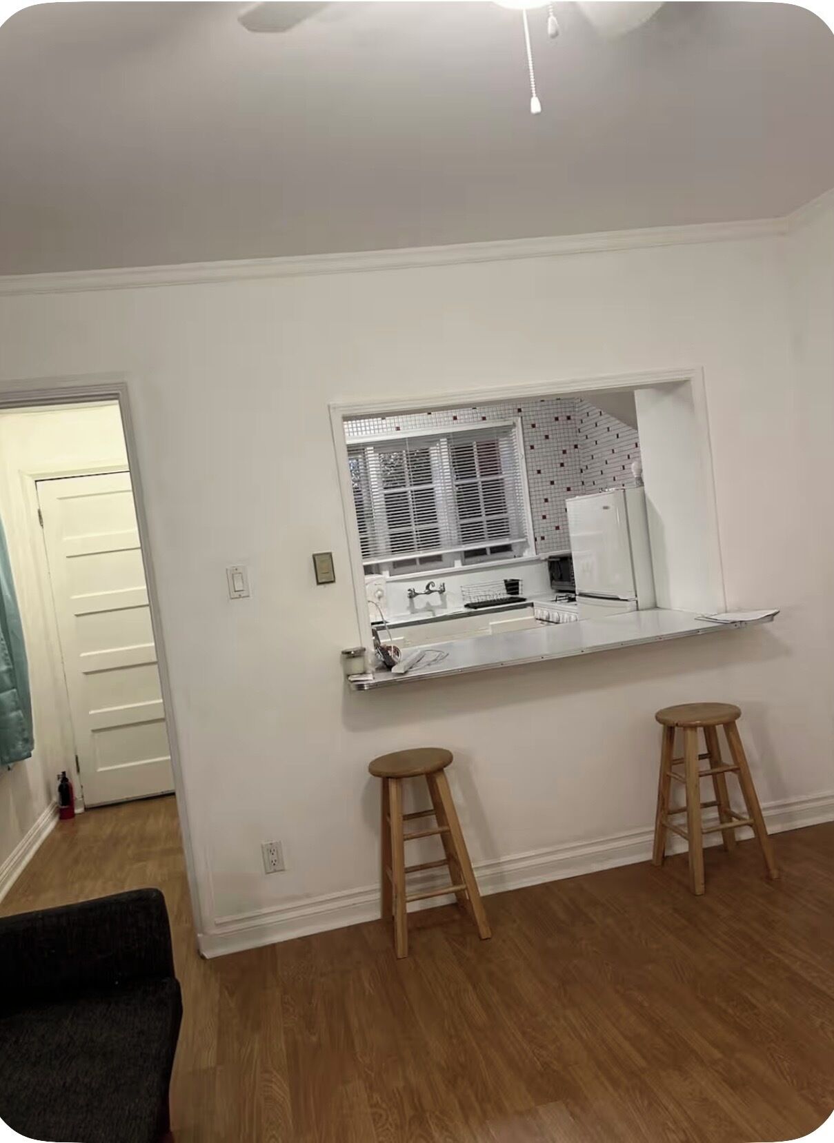 Una habitación con una pared blanca, una ventana con persianas, una encimera blanca, dos taburetes de madera y un ventilador de techo.