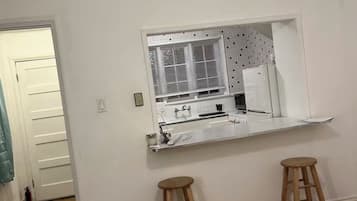 Una habitación con una pared blanca, una ventana con persianas, una encimera blanca, dos taburetes de madera y un ventilador de techo.