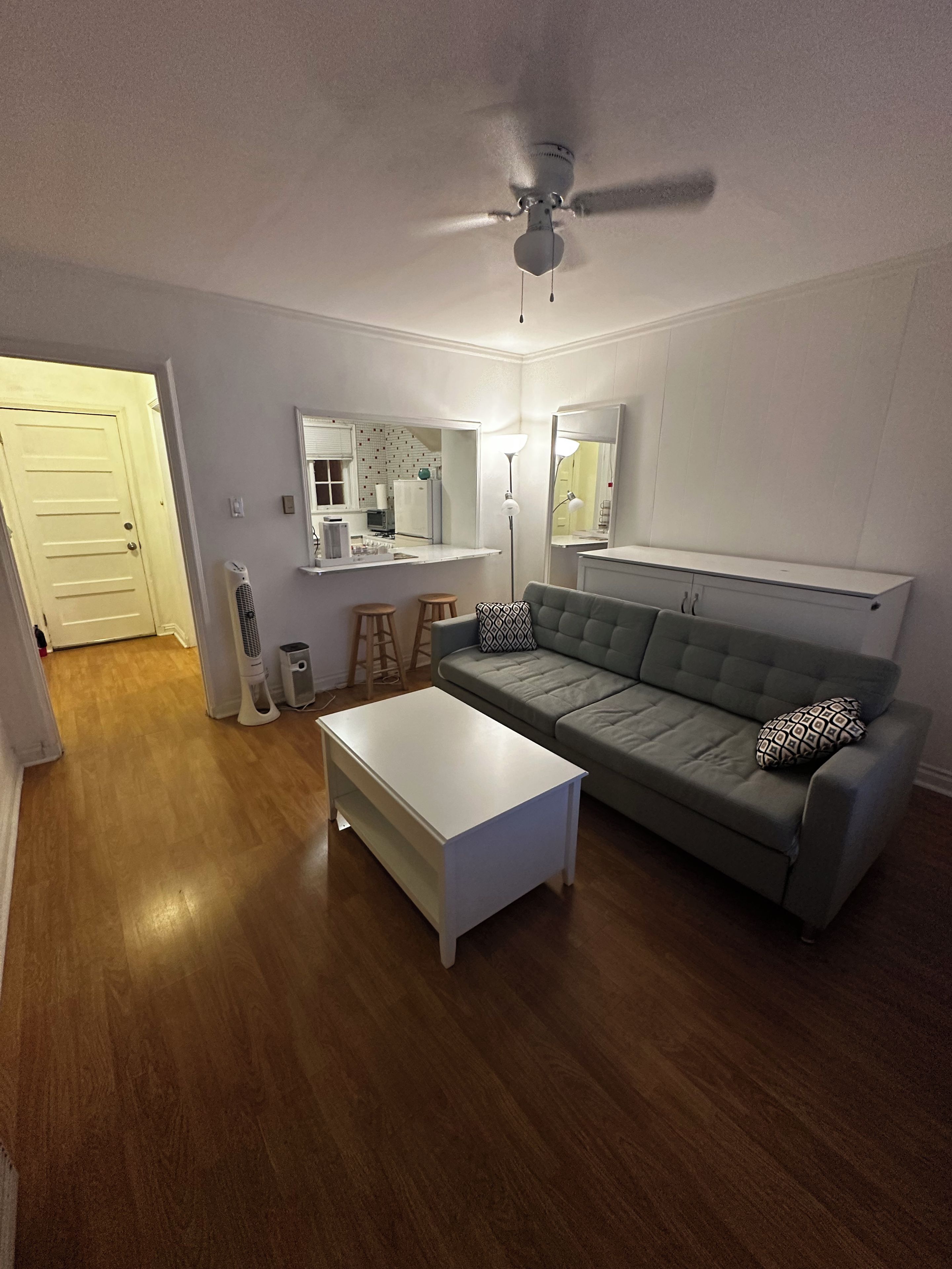 A living room with a grey sofa, a white coffee table, a ceiling fan, and a kitchen area in the background.