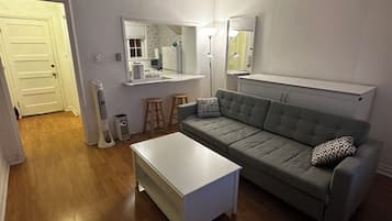 A living room with a grey sofa, a white coffee table, a ceiling fan, and a kitchen area in the background.