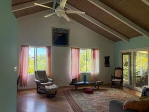 A spacious living room with a high ceiling, large windows, a ceiling fan, and a view of greenery outside.