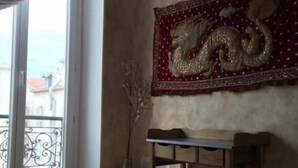 A room with a wooden floor, a table, a vase with branches, and a wall tapestry with a dragon design.