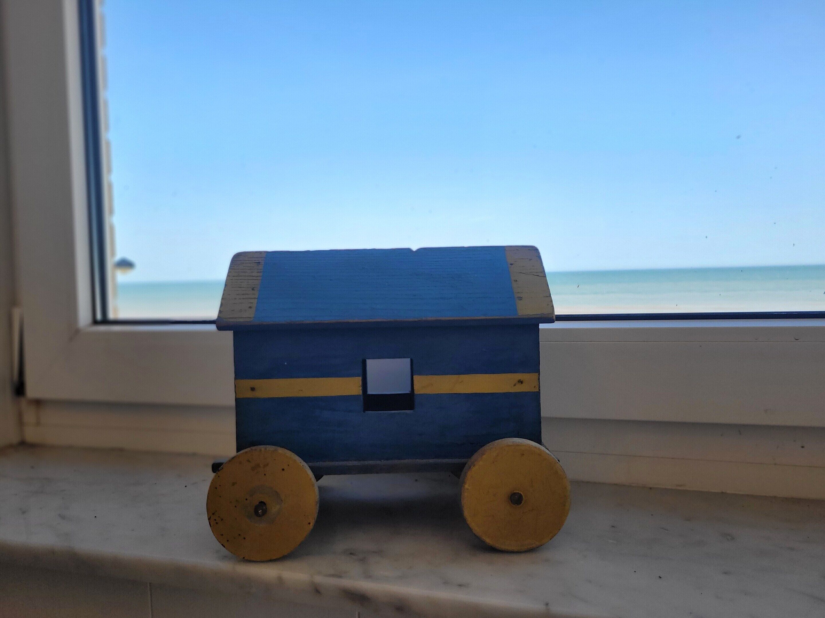 Eine kleine, auf Rädern stehende Holzhütte, platziert auf einem Vorsprung neben einem Fenster mit Blick auf einen Strand.