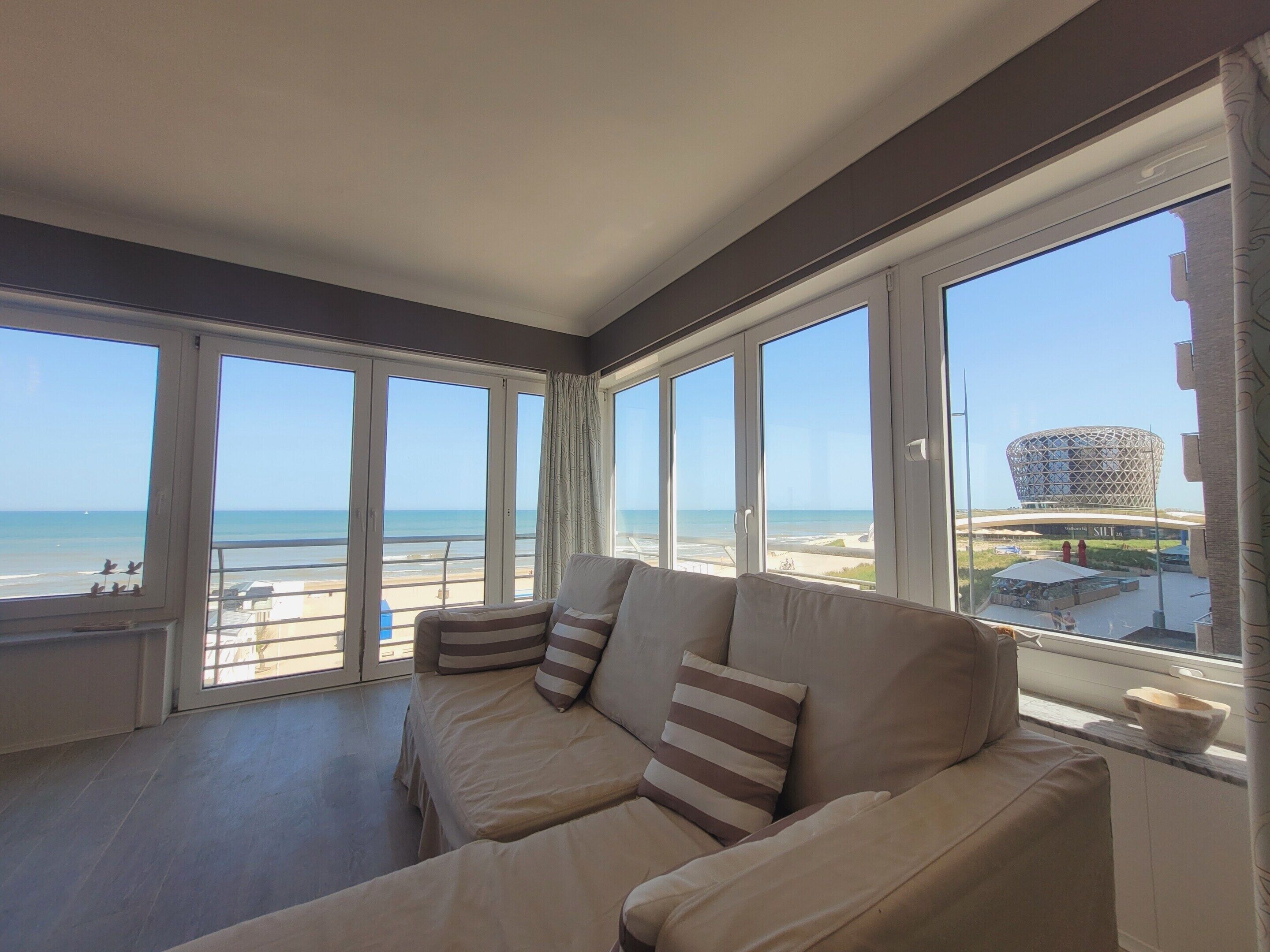 Ein Zimmer mit einer Couch, die zu großen Fenstern mit Blick auf einen Strand und ein markantes Gebäude hinweist.