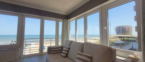 A room with a sofa facing large windows overlooking a beach and a distinctive building.