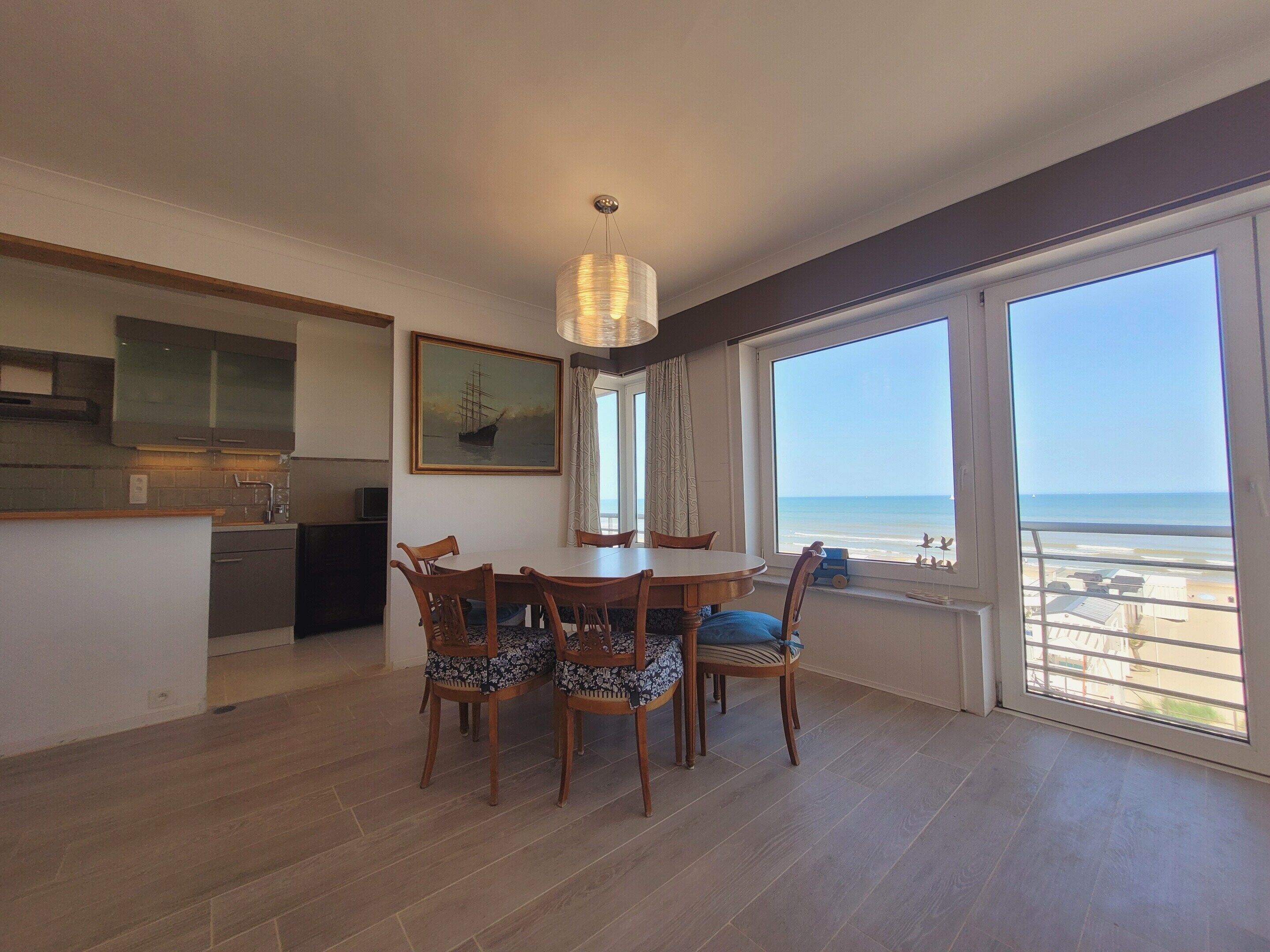 A dining area with a table and chairs, a kitchen in the background, and a large window with a view of the sea.