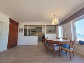 A dining area with a round table and chairs, a kitchen in the background, and a large window with a view. - Apartment With Panoramic Sea Views (Middelkerke)