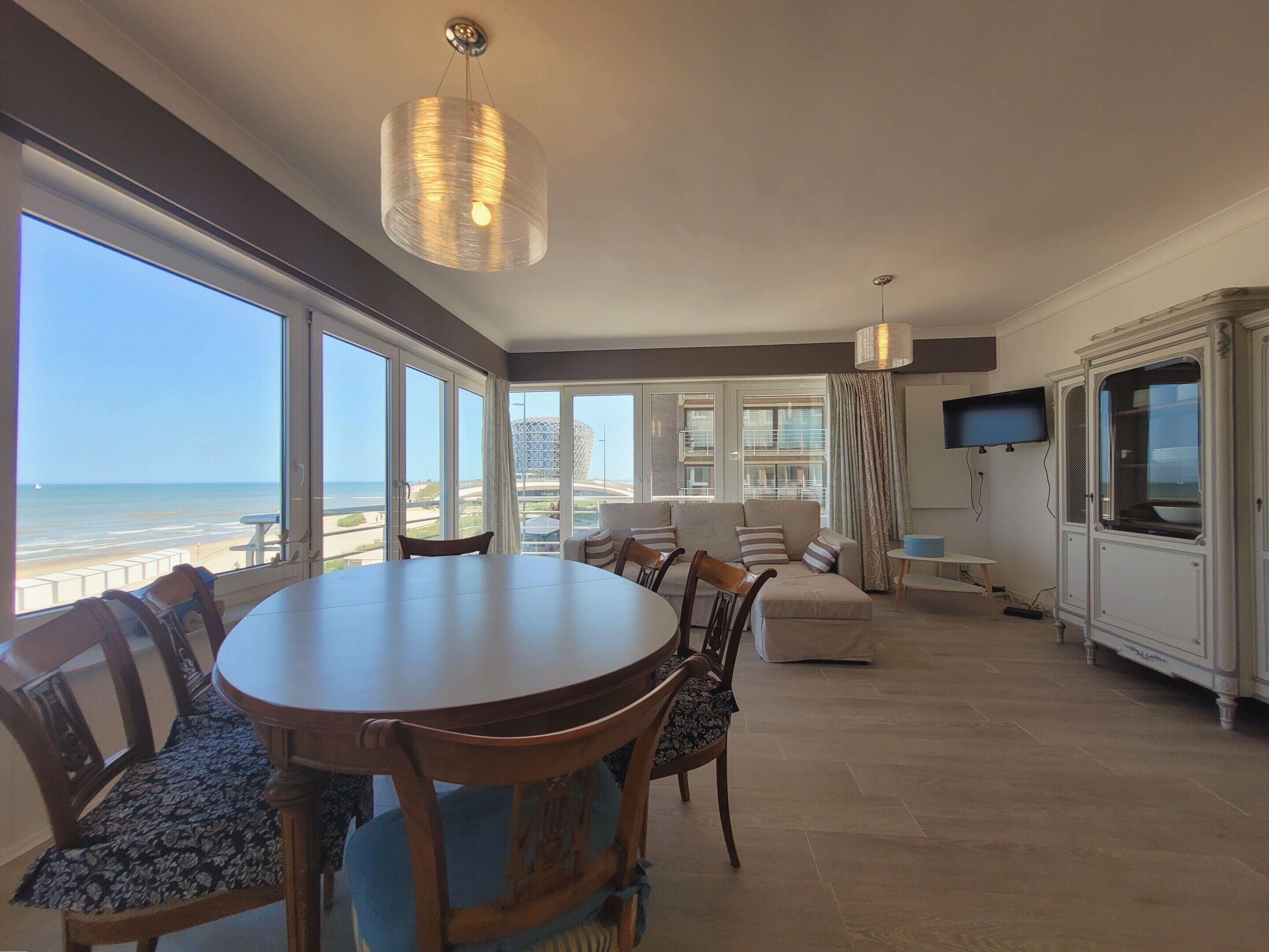 A spacious dining area with a large table and chairs, a television mounted on the wall, and a view of the beach through large windows.